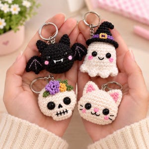 May include: Four crochet keychains featuring a black bat with white fangs, a white ghost wearing a purple-banded black witch hat, a white skull adorned with purple and yellow flowers, and a cream-colored cat with pink inner ears and whiskers.