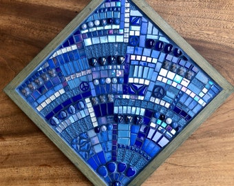 May include: A mosaic artwork in a diamond-shaped wooden frame. The mosaic features a variety of blue tiles, including squares, rectangles, and rounded pieces. The design incorporates peace symbols and a radial pattern, creating a visually appealing piece.