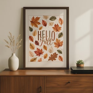 May include: A framed wall art print featuring the text "HELLO Fall" in brown lettering, surrounded by a variety of illustrated autumn leaves in shades of orange, red, yellow, brown, and green against a cream-coloured background.