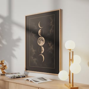 May include: A framed vertical wall art print featuring a sequence of five moon phases in varying illumination against a dark, cloudy background. The artwork is enclosed in a light-toned wooden frame and displayed on a wooden surface next to a lamp.