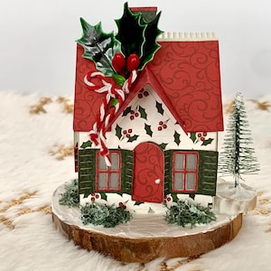 May include: A festive Christmas house ornament with a red roof and shutters, decorated with holly and berries. The house sits on a wooden base with a small Christmas tree and greenery, creating a charming holiday scene.