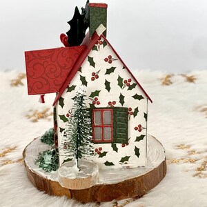 May include: A decorative Christmas house figurine with a red roof and chimney, holly berry and leaf pattern, and a small Christmas tree. The house is set on a wooden base.