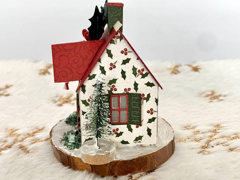 May include: A decorative Christmas house figurine with a red roof and chimney, holly berry and leaf pattern, and a small Christmas tree. The house is set on a wooden base.