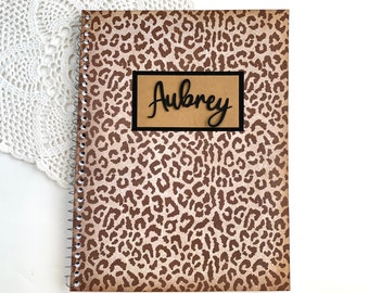 May include: A brown spiral notebook with a leopard print cover. The notebook has a brown paper label with the name "Aubrey" written in black cursive.