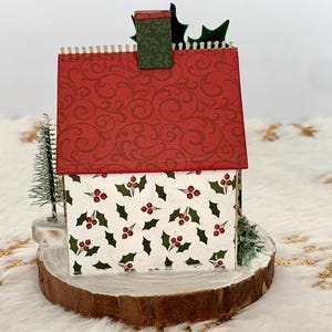 May include: A miniature Christmas house with a red roof featuring a swirling pattern. The house walls are white with a holly berry design. It sits on a wooden base with small evergreen trees and holly accents.