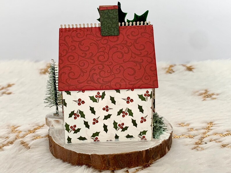 May include: A miniature Christmas house with a red roof featuring a swirling pattern. The house walls are white with a holly berry design. It sits on a wooden base with small evergreen trees and holly accents.