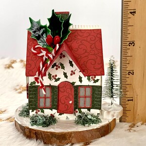 May include: A miniature Christmas house with a red roof and green shutters. The house is decorated with holly and berries, and sits on a wooden base. A small Christmas tree is next to the house. The house is approximately 3 inches tall.
