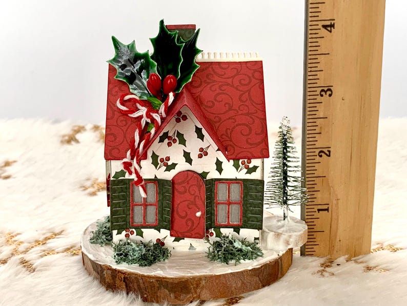 May include: A miniature Christmas house with a red roof and green shutters. The house is decorated with holly and berries, and sits on a wooden base. A small Christmas tree is next to the house. The house is approximately 3 inches tall.