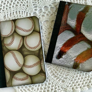 May include: Two small notebooks with different designs. One notebook has a baseball pattern with white baseballs on a brown background. The other notebook has a white and orange abstract pattern.