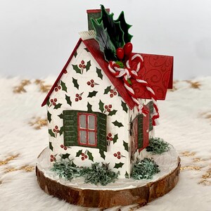 May include: A decorative Christmas house figurine. The white house features a red roof, green shutters, and is decorated with holly and berry patterns. A chimney, holly, and candy cane accents complete the festive design. The house sits on a wooden base.