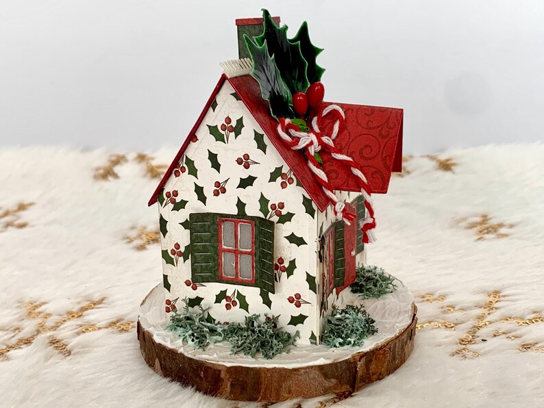 May include: A decorative Christmas house figurine. The white house features a red roof, green shutters, and is decorated with holly and berry patterns. A chimney, holly, and candy cane accents complete the festive design. The house sits on a wooden base.