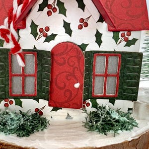 May include: A miniature Christmas house with a red roof and door, green shutters, and holly berry accents. The house sits on a wooden base with faux snow and greenery, and a small Christmas tree is visible in the background.