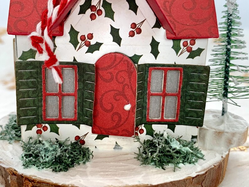 May include: A miniature Christmas house with a red roof and door, green shutters, and holly berry accents. The house sits on a wooden base with faux snow and greenery, and a small Christmas tree is visible in the background.