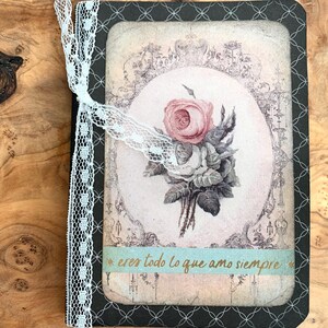 May include: A vintage-style journal with a floral design. The cover features a bouquet of roses in shades of pink and white, framed by an ornate border. A light blue ribbon with the text "eres todo lo que amo siempre" is attached.