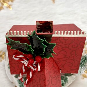May include: A red Christmas-themed decorative house with a chimney, adorned with holly leaves, red berries, and a candy cane bow. The house has a white trim and a patterned design.