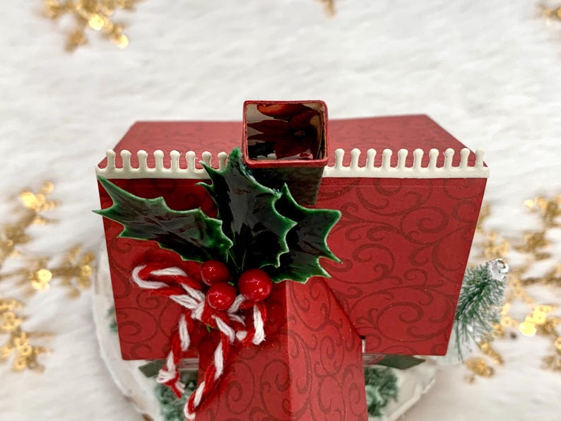 May include: A red Christmas-themed decorative house with a chimney, adorned with holly leaves, red berries, and a candy cane bow. The house has a white trim and a patterned design.
