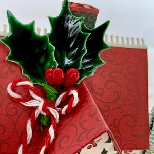 May include: A festive Christmas-themed miniature house with a red patterned exterior. The house is adorned with green holly leaves, red berries, and a candy cane-style bow. A small chimney and decorative trim complete the design.