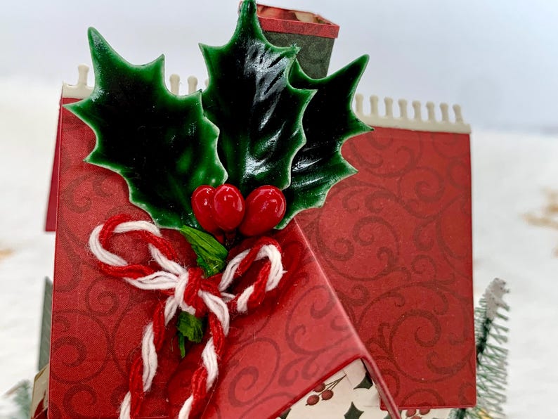 May include: A festive Christmas-themed miniature house with a red patterned exterior. The house is adorned with green holly leaves, red berries, and a candy cane-style bow. A small chimney and decorative trim complete the design.