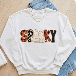 以下が含まれることがあります： 胸元に「SPOOKY」の文字がプリントされた白いクルーネックのスウェットシャツ。文字はオレンジと黒のバッファローチェック、白い花柄の黒地、そしてクリーム色の2体の幽霊のイラストなど、さまざまなパターンで塗りつぶされています。