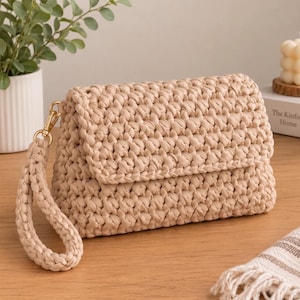 Könnte beinhalten: Eine beige gehäkelte Clutch mit einem dicken, strukturierten Maschenmuster und einer passenden Handschlaufe, die mit einem goldfarbenen Metallverschluss befestigt ist. Die rechteckige Tasche hat einen Umschlagdeckel aus kordelartigem Garn.