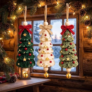 May include: Three hanging crochet Christmas tree ornaments. The left and right trees are dark and light green, while the center tree is cream. Each features red and gold beads, gold star accents, a top bow, a wooden bead, and a gold jingle bell.