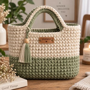 Può includere: Un cestino all'uncinetto bicolore con una sezione superiore color crema e una sezione inferiore verde salvia. Presenta due manici integrati, una nappa decorativa con una perla di legno e un'etichetta in similpelle marrone con la scritta HANDMADE e un cuore.