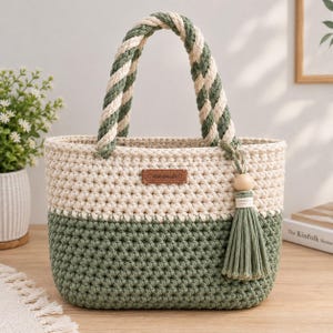 Può includere: Una borsa tote all'uncinetto bicolore con una sezione superiore color crema e una sezione inferiore verde salvia. La borsa presenta spessi manici in corda intrecciata color crema e verde salvia, un'etichetta in pelle marrone con scritto Handmade e una nappa verde salvia.
