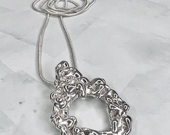May include: A silver necklace with a large, abstract, textured pendant. The pendant is shaped like a circle with an open center. The chain is a thin, silver snake chain.
