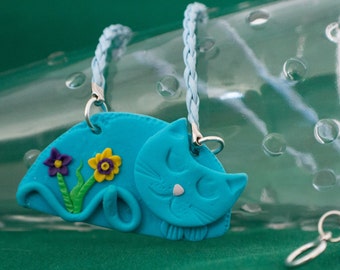 May include: A blue cat charm with yellow and purple flowers, attached to a light blue braided cord necklace.