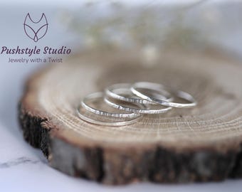 May include: Four silver rings with textured surfaces are displayed on a rustic wooden slice. The rings are stacked together, showcasing their delicate design. The rings are from Pushstyle Studio, Jewelry with a Twist.