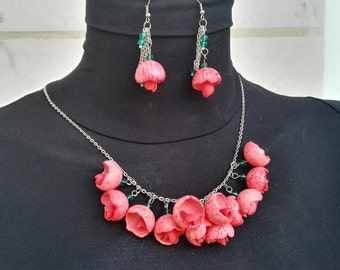May include: A coral-colored necklace and earring set. The necklace features a silver chain with coral-colored flower-shaped pendants. The earrings have a similar design, with small green beads. The jewelry is displayed on a black mannequin.