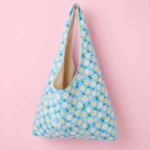 May include: A triangular hobo bag featuring a light blue background with a repeating pattern of white daisies with yellow centres and small green leaves. The interior of the bag is lined with a solid beige fabric, visible through the open top.