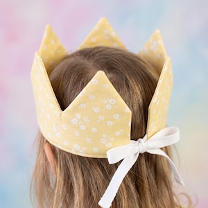Puede incluir: Una corona de tela amarilla con un pequeño estampado floral blanco y cinco puntas. La corona se asegura en la parte posterior con una cinta tejida blanca atada en un lazo, lo que permite un ajuste regulable al contorno de la cabeza.