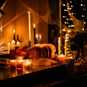 Puede incluir: Una gran calabaza naranja sirve como centro de mesa, rodeada de m&uacute;ltiples velas encendidas de diversas alturas y colores, incluyendo cirios blancos y negros y peque&ntilde;as velas votivas. Telara&ntilde;as artificiales cubren la escena con una luz c&aacute;lida.