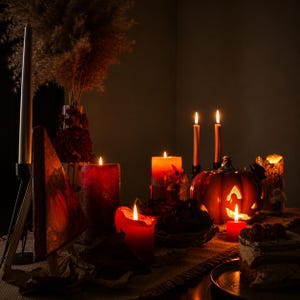 Puede incluir: Un arreglo festivo que incluye una linterna de calabaza naranja tallada, varias velas de pilar rojas y doradas con llamas parpadeantes, una peque&ntilde;a pintura triangular de una calabaza, arreglos florales secos y un trozo de pastel.