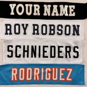 May include: Four rectangular fabric patches with embroidered lettering. The top patch is black with white text reading YOUR NAME. The second is white with dark blue text reading ROY ROBSON. The third is white with black text reading SCHNIEDERS. The bottom is blue with orange text reading RODRIGUEZ.