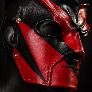 May include: A black and red leather mask with a striking design. The mask features a black base with bold red accents, including angular shapes and a prominent facial design. Gold-coloured rivets secure the straps.