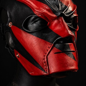 May include: A black and red leather mask with a striking design. The mask has a black base with bold red stripes and accents, including a prominent nose and eye openings. The mask is likely for cosplay or costume use.