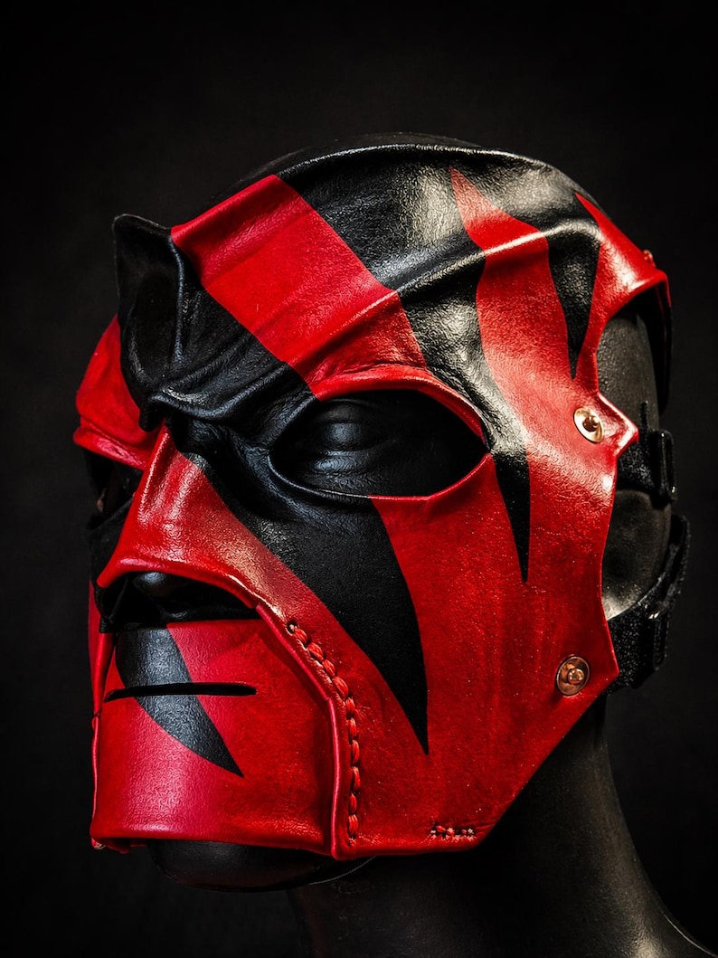 May include: A black and red leather mask with a striking design. The mask has eye and mouth openings, and is secured with straps and rivets. The mask is displayed on a black mannequin head. The mask's design features bold, angular patterns.