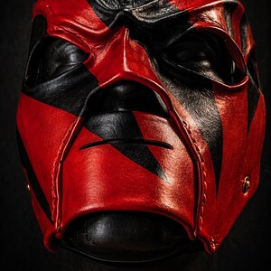 May include: A red and black mask with a striking design. The mask is crafted from red and black leather, with a stitched seam and eye openings. The mask is designed for wear and is likely for costume or cosplay purposes.