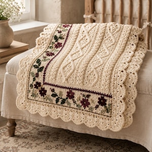 Peut inclure: Une couverture crochetée de couleur crème présentant un panneau central avec des motifs de torsades et de losanges complexes. La bordure est ornée d'un motif floral de fleurs rouge foncé et de feuilles vertes, avec un bord festonné.