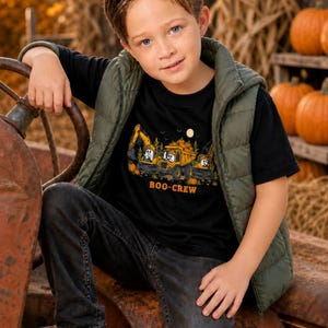 May include: A black short-sleeved t-shirt featuring a graphic of construction vehicles driven by ghosts, with pumpkins and the text "BOO-CREW" printed in orange. The shirt is worn under an olive green quilted vest, paired with dark gray jeans.