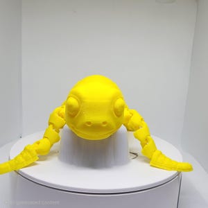 Pode incluir: Uma figura de tartaruga articulada impressa em 3D de cor amarela brilhante, com uma carapaça lisa e arredondada e membros segmentados que se estendem para fora. A criatura apresenta uma cabeça distinta com duas pequenas narinas e olhos fechados.