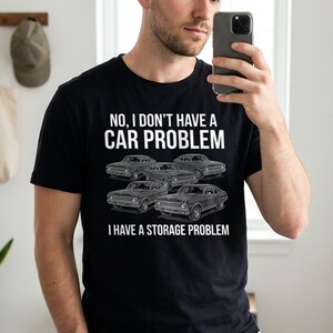 Puede incluir: Una camiseta negra con texto en blanco que dice: NO, I DON'T HAVE A CAR PROBLEM, I HAVE A STORAGE PROBLEM. El diseño incluye una ilustración gráfica de cuatro coches deportivos antiguos dispuestos en un grupo en el centro de la prenda.