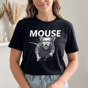 Może przedstawiać: Czarny t-shirt z krótkim rękawem z nadrukiem graficznym przedstawiającym mysz z ludzką twarzą. Słowo MOUSE jest wydrukowane dużymi białymi wielkimi literami nad ilustracją. Wzór zawiera cienkie białe linie rozchodzące się promieniście ze środka.