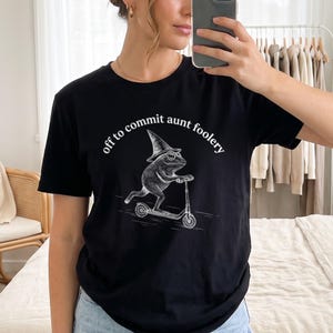 Puede incluir: Una camiseta negra de manga corta con un gráfico blanco de una rana con sombrero puntiagudo montada en un patinete. El texto sobre la rana dice "off to commit aunt foolery" en una fuente blanca curva. Tiene cuello redondo.