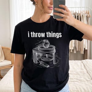 以下が含まれることがあります： 黒の半袖Tシャツ。前面中央に「i throw things」という白い文字と、足踏みペダル付きのろくろで粘土の壺を成形している人物のグラフィックイラストがプリントされています。