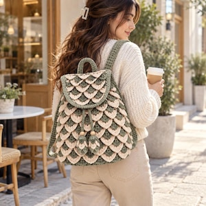 Puede incluir: Una mochila de ganchillo con un patrón de escamas superpuestas en hilo verde salvia y crema alternados. La bolsa incluye un asa superior, dos correas para los hombros, un cierre de cordón con dos borlas decorativas y una solapa redondeada.