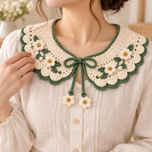 Op de afbeelding: Een crèmekleurige gehaakte kraag met een geschulpte rand afgezet in donkergroen. De kraag is versierd met kleine witte bloemen met gele harten en groene bladeren. Twee lange groene linten hangen vanuit het midden, elk eindigend in een bloem.