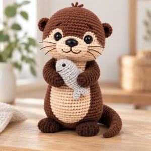 May include: A crocheted otter stuffed animal made with dark brown yarn for the body and light beige yarn for the face and belly. The otter has black plastic eyes, a black nose, and embroidered whiskers, and it is holding a small gray fish.